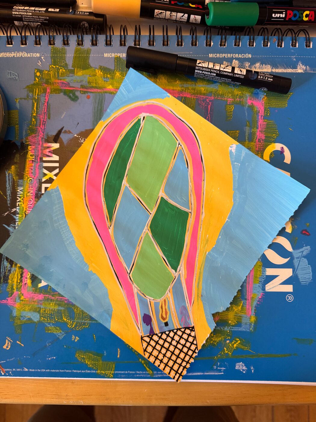 A hot-air balloon painted with a pink outline and green and blue panels over a black crosshatched basket, on a yellow shape laid over a light blue sheet with a torn edge