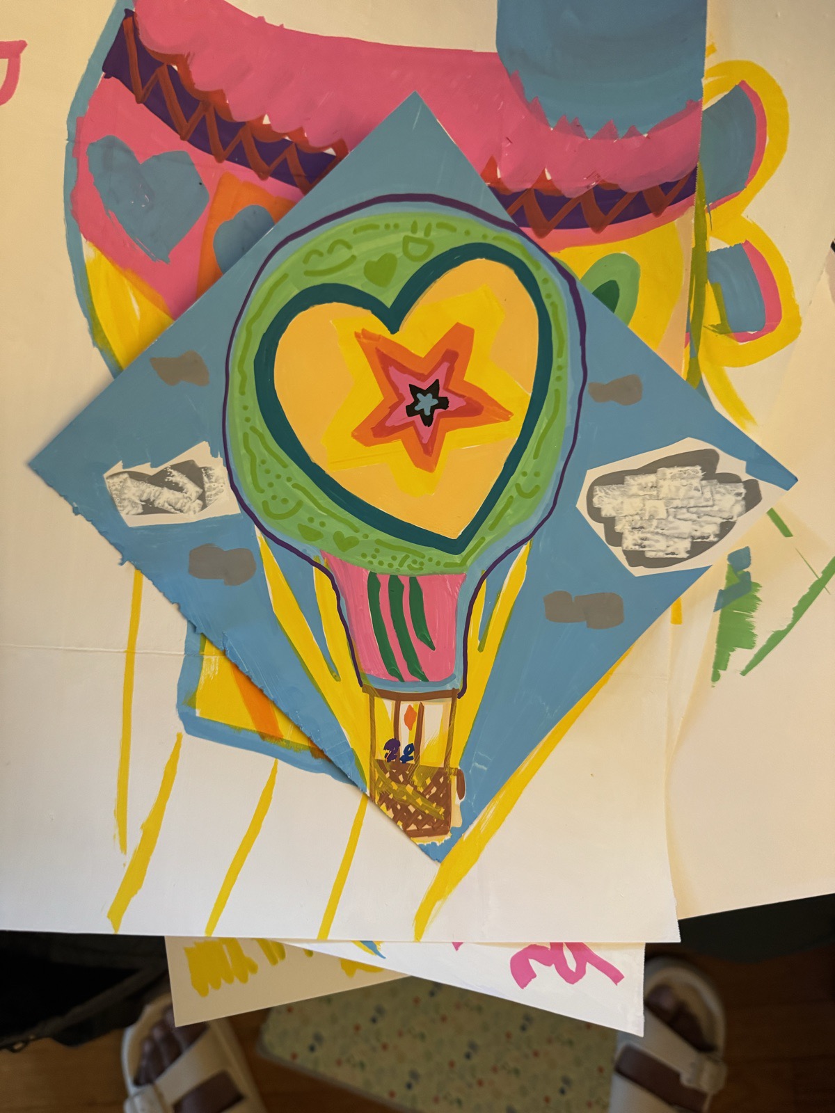 Hot-air balloon with a heart and a star inside it, painted in green, yellow, pink and orange on a blue diamond of paper, with grayscale photo cutouts as clouds, laid over a larger painted sheet