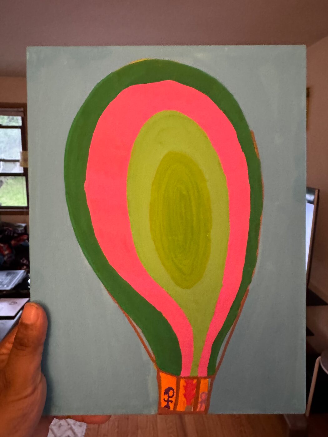 Painting on canvas board: a large hot-air balloon in dark green, pink and light green bands around an olive center, on a gray-blue ground, with small figures in its orange basket
