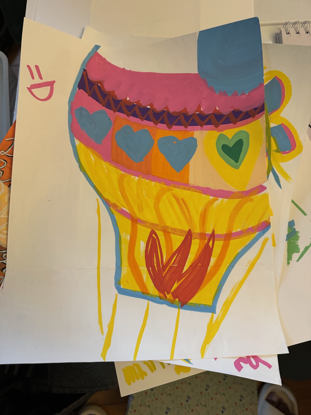 Large balloon painted in yellow and orange with a pink and purple zigzag band, blue and green hearts, and a red flame shape at the base, on a white sheet