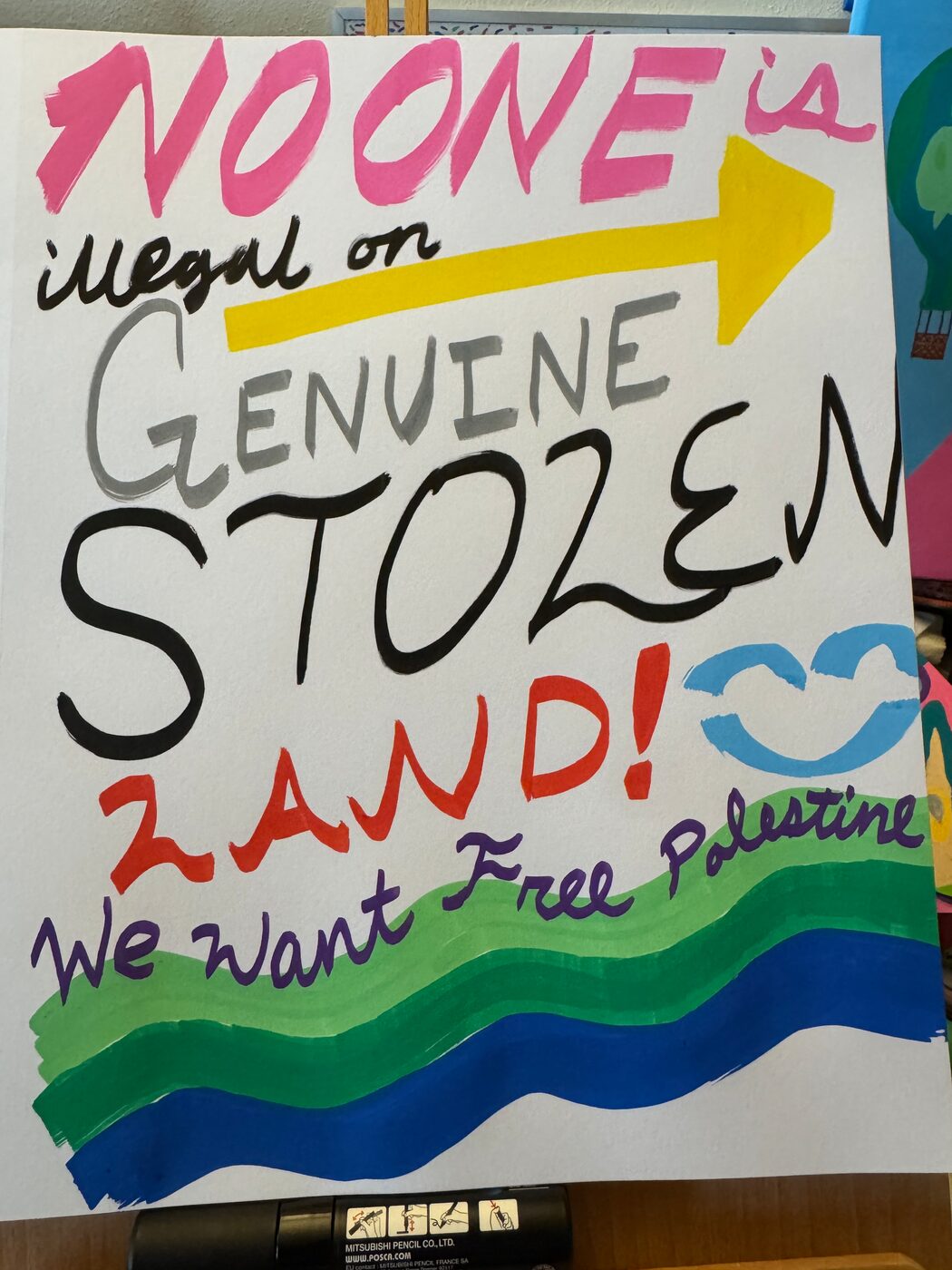 A sign in marker and paint: NO ONE is illegal on GENUINE STOLEN LAND!, with a yellow arrow and a light blue smiling face, and We want Free Palestine written above green and blue waves