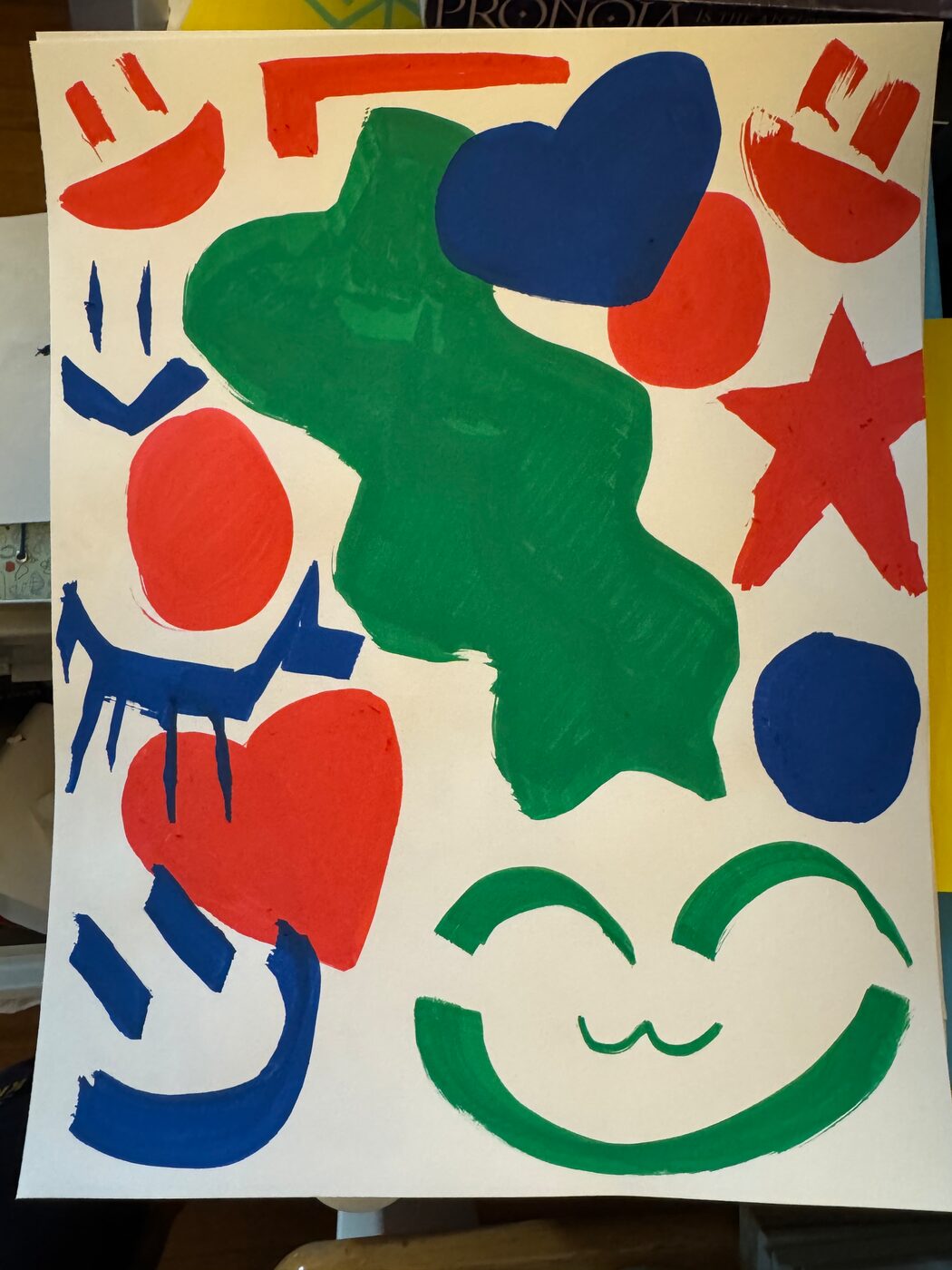 Painted shapes in red, dark blue and green on a cream sheet: a large green blob, hearts, circles, a red star, and five smiling faces, the biggest a green one with a cat's mouth