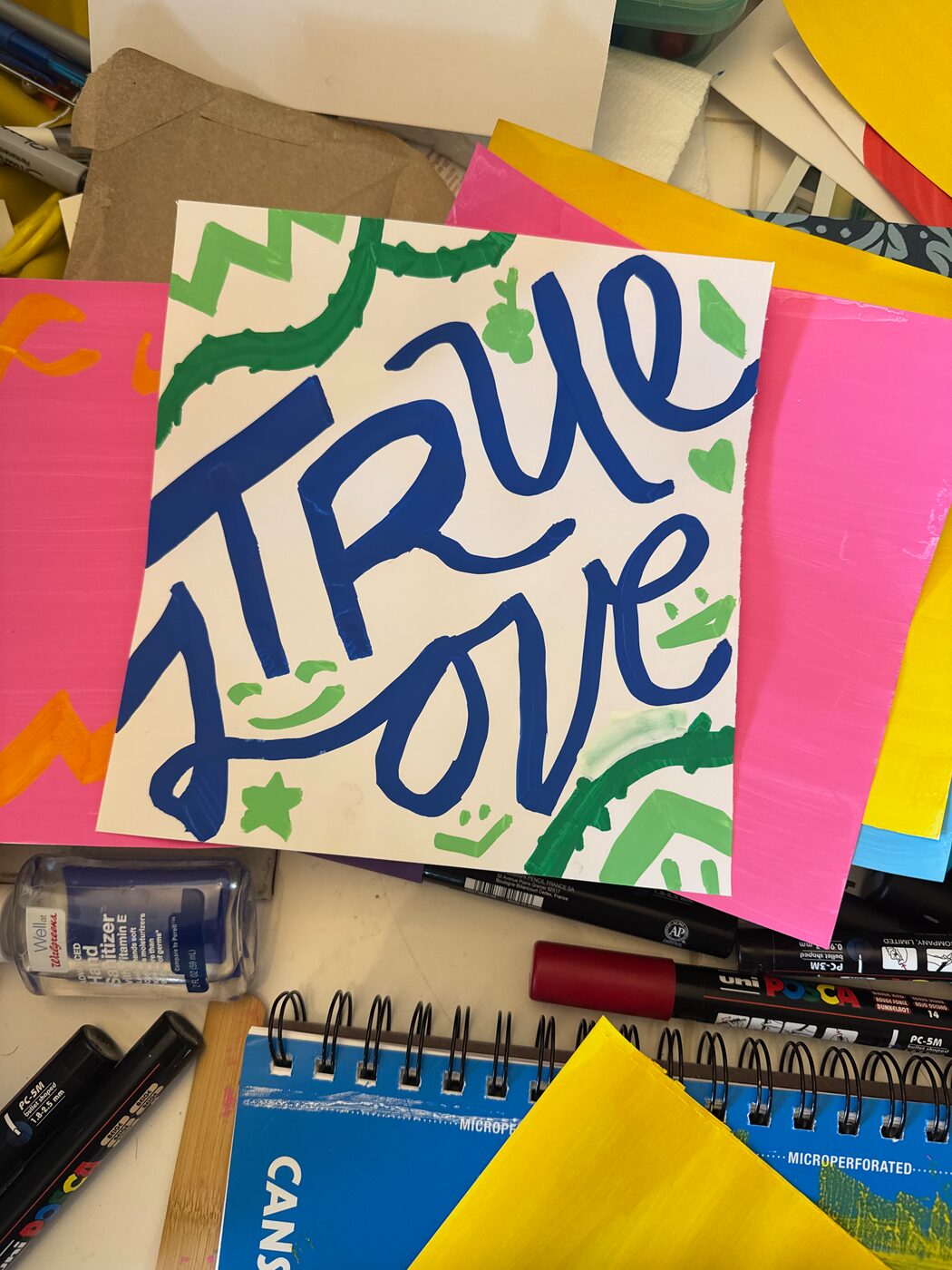 TRUE LOVE in blue brush script on a white sheet with green zigzags, smiling faces, a star and a heart, laid on pink and yellow paper
