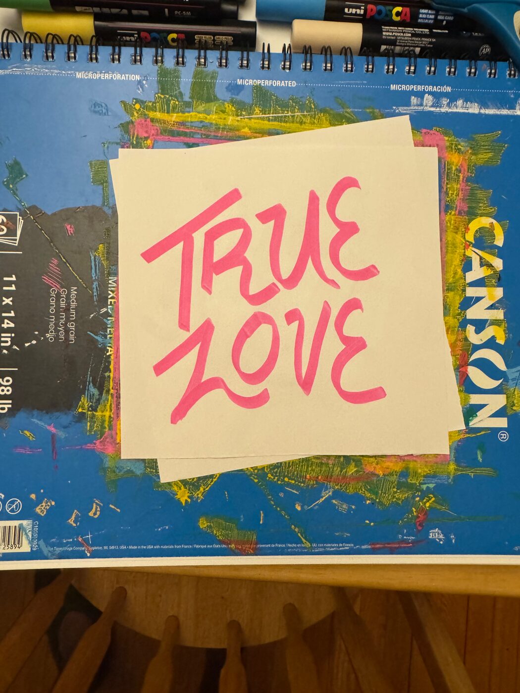 TRUE LOVE in pink marker capitals on a white sheet laid over a paint-streaked sketchbook
